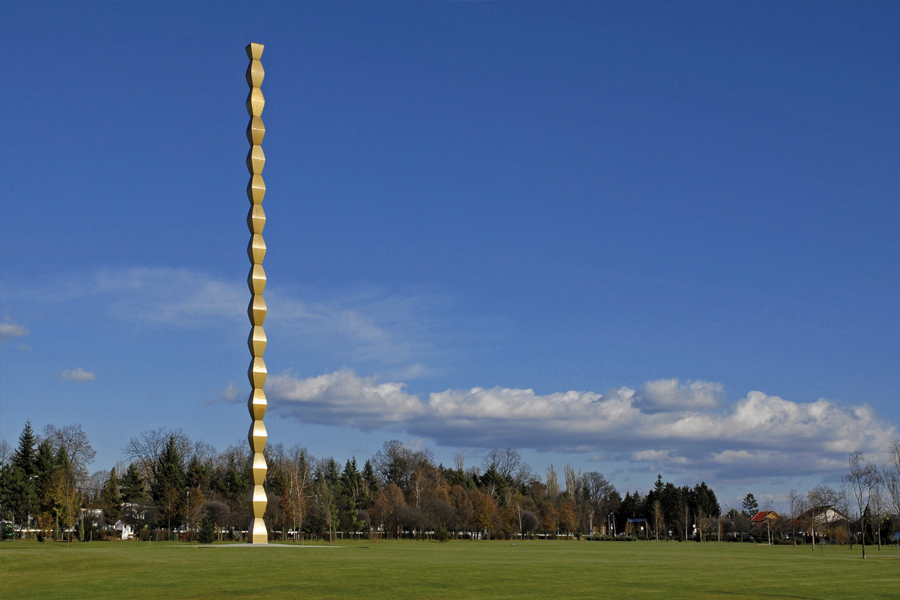 The Endless Column: A tall, modular golden-bronze sculpture stretching toward a clear blue sky in an open green park.