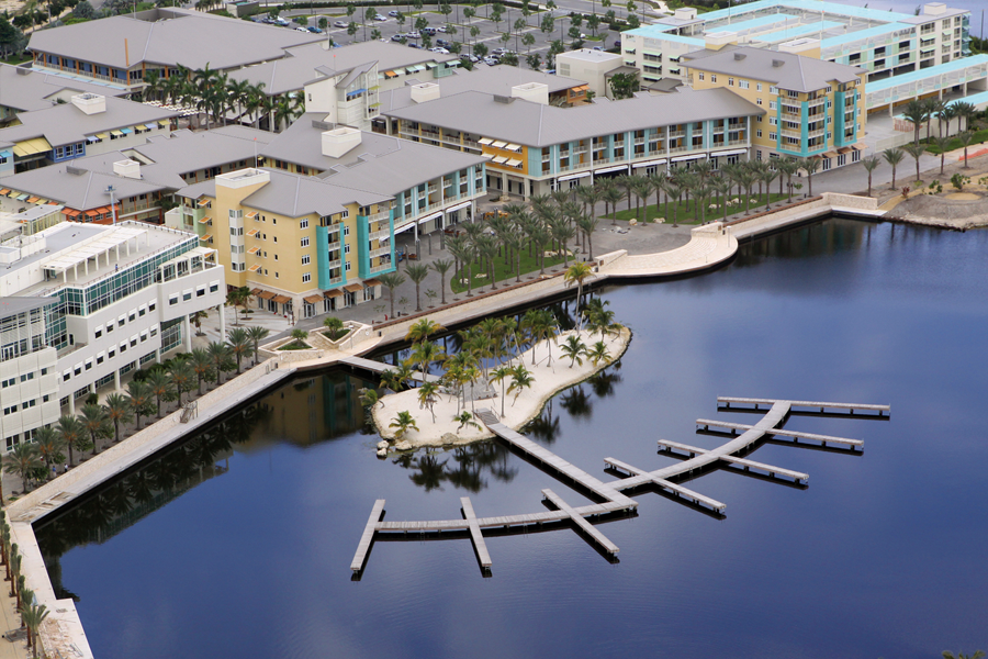 Aerial view of a modern waterfront development featuring multi-story buildings, a circular harbor with a wooden pier, and a small palm-covered island.