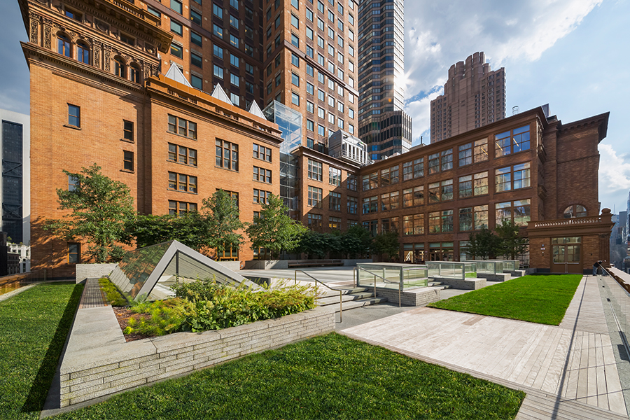 A daytime view of a modern rooftop terrace featuring green lawns, stone walkways, and a triangular glass skylight, set against a backdrop of historic red-brick buildings and city skyscrapers.