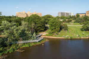A high-angle view of an elevated walkway curving along the Schuylkill River. People lounge on a grassy hill near the Philadelphia Museum of Art, with apartment buildings in the distance.
