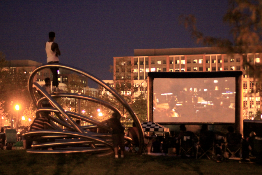 A nighttime scene of an outdoor movie screening in a park. Audience members sit in lawn chairs and a circular metal sculpture facing a large inflatable screen, against a backdrop of lit office buildings.