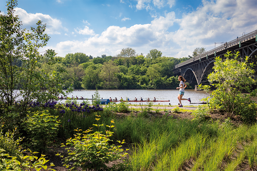 A woman jogs along a paved riverfront path lined with purple irises and greenery, while a rowing shell passes by on the water near a large arched bridge.