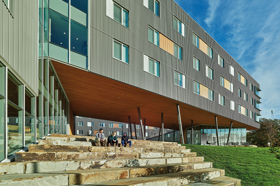 Three students sit on a wide, tiered stone staircase under the wooden soffit of a large building cantilever. The facade above shows textured grey metal siding punctuated by white and orange window accents.