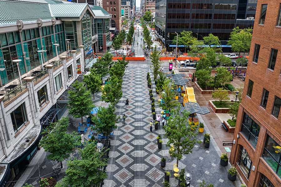 High-angle view of a wide pedestrian mall lined with trees, outdoor seating, and modern buildings, featuring a distinctive gray and white diamond-patterned stone pavement.