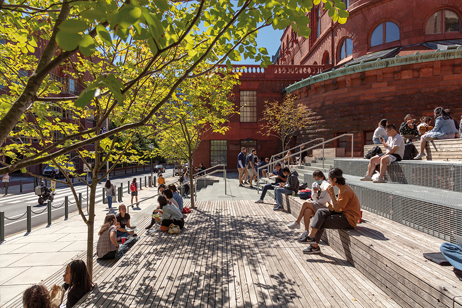 A ground-level perspective of the tiered wooden seating area filled with students eating and talking. The scene is framed by leafy green trees and the ornate red brick and terra cotta architecture of the campus.