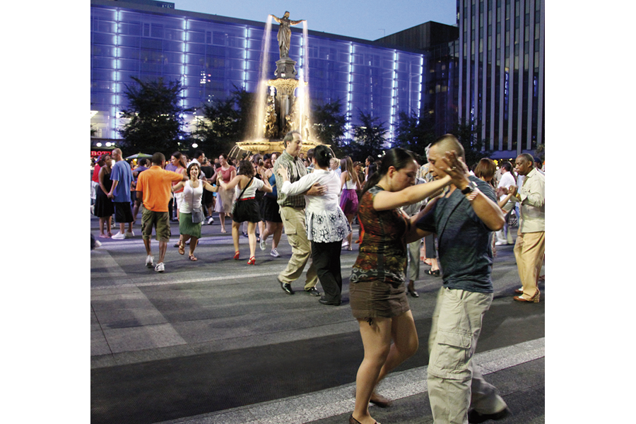 A crowd of people partner dancing in a city square at night, with a glowing fountain and illuminated blue building in the background.