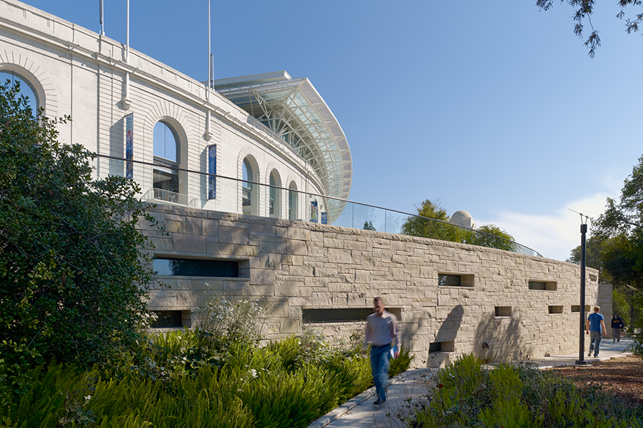 A view of a textured stone retaining wall with horizontal windows, situated below the arched facade of the California Memorial Stadium.