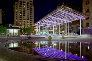 A nighttime view of the plaza featuring the glass pavilion illuminated with purple lights, reflected in the still water of the circular fountain.