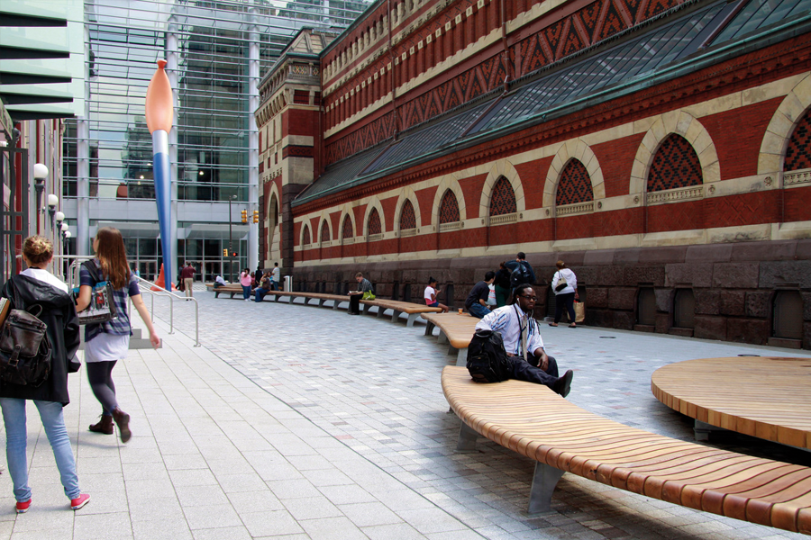 A wide pedestrian walkway with curved wooden benches running alongside a historic red brick facade.
