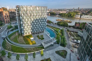 High-angle aerial view of the U.S. Embassy in London, showing its glass cube structure with ETFE plastic sails, a semi-circular moat, and landscaped gardens along the River Thames.