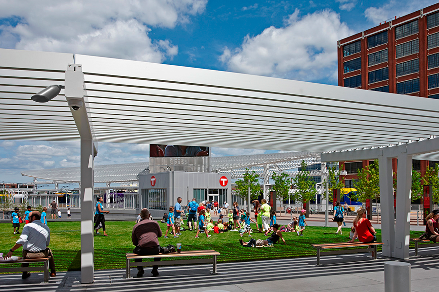 A sunlit view from beneath a white louvered pergola looking out onto a green lawn where children play and people sit on benches near a transit entrance.