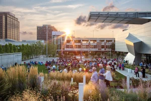 A sunset view of a crowd gathered on the garden lawn for an outdoor performance, with the modern steel museum cantilever and city skyline in the background.
