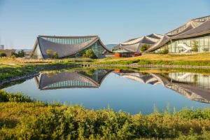A scenic view of the tent-like campus buildings reflected in a calm pond, surrounded by green meadow grass under a clear blue sky.