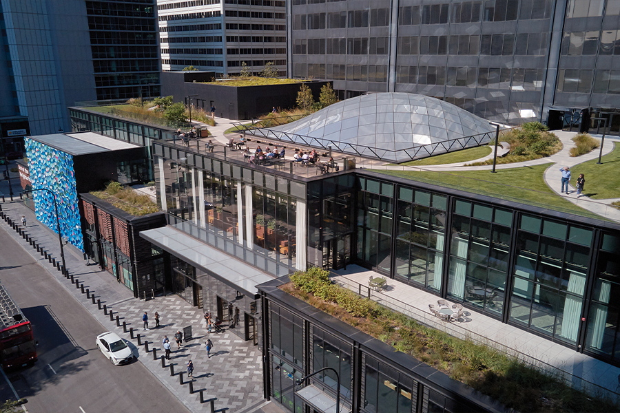 An elevated view of a multi-level modern building featuring extensive rooftop greenery, an outdoor dining deck, and a prominent glass dome skylight.