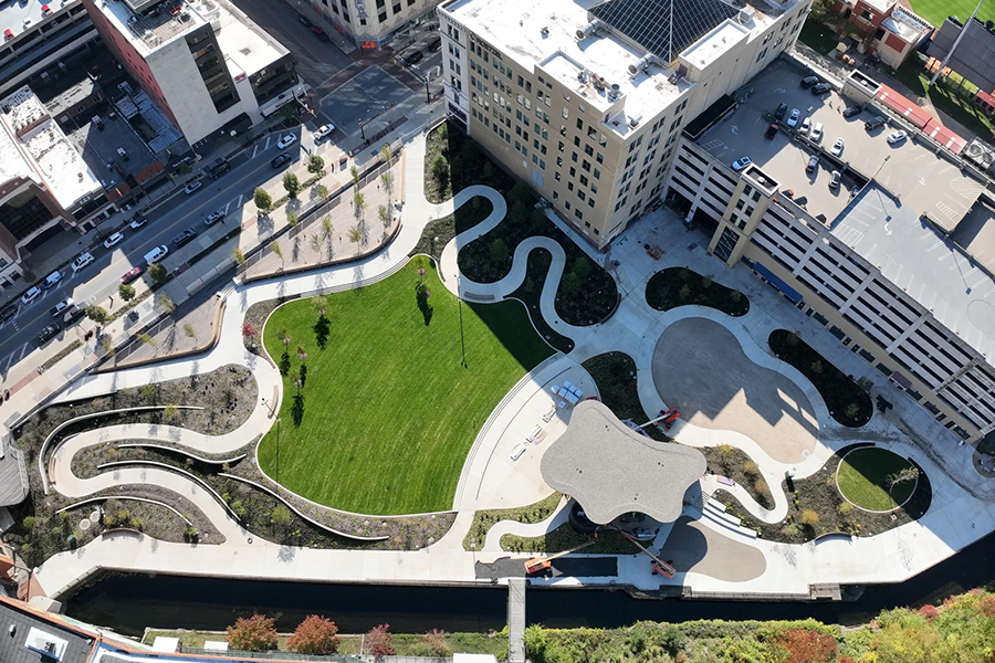 Aerial view of a completed urban park with a large green lawn, curved concrete paths, and a modern performance stage.