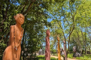 A collection of tall, stylized clay or stone sculptures of human figures with beaded necklaces standing in a sunny, wooded park.