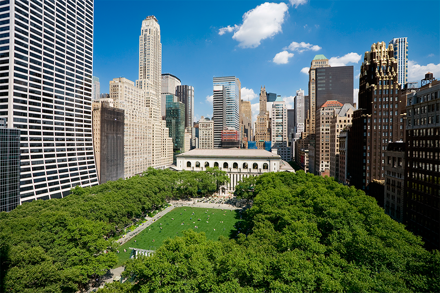 A high-angle view of Bryant Park's green lawn and the New York Public Library, framed by dense green trees and a backdrop of various Midtown Manhattan skyscrapers.