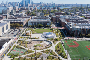 Wide aerial shot of a city park with a skyline in the background, showing a baseball field, basketball court, and circular central building.