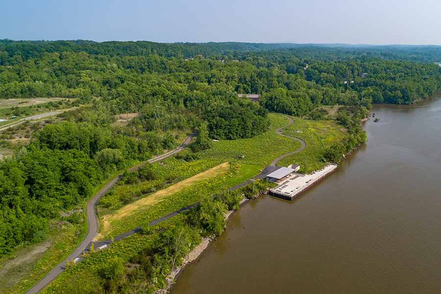 Aerial view of a riverside park featuring a modern pier, a sheltered pavilion, and winding paved trails through lush green meadows and dense forest.