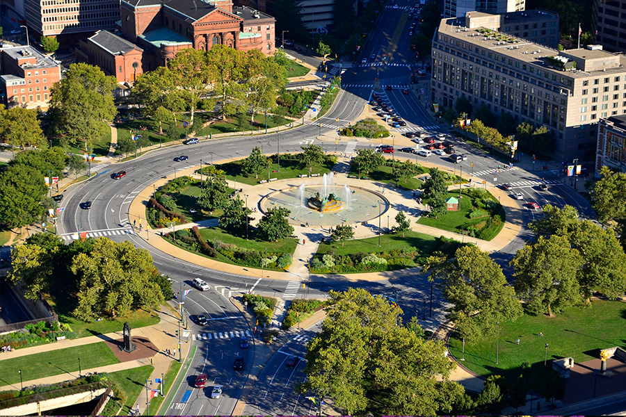 Aerial view of a large urban traffic circle park with a central fountain and surrounding green space.
