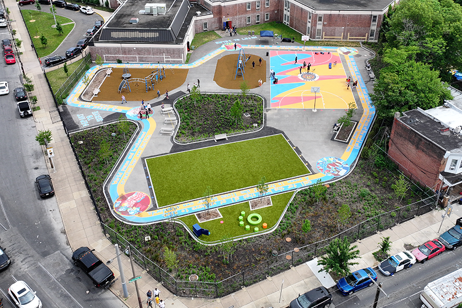 An aerial view of a vibrant, newly renovated schoolyard featuring a colorful basketball court, a playground with woodchips, a small turf field, and paved walking paths winding through green garden beds.