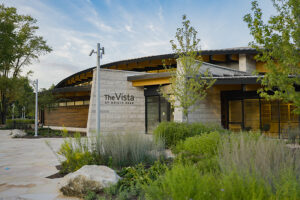 Exterior ground-level view of The Vista at Origin Park building featuring stone masonry, wood paneling, and landscaped garden beds.