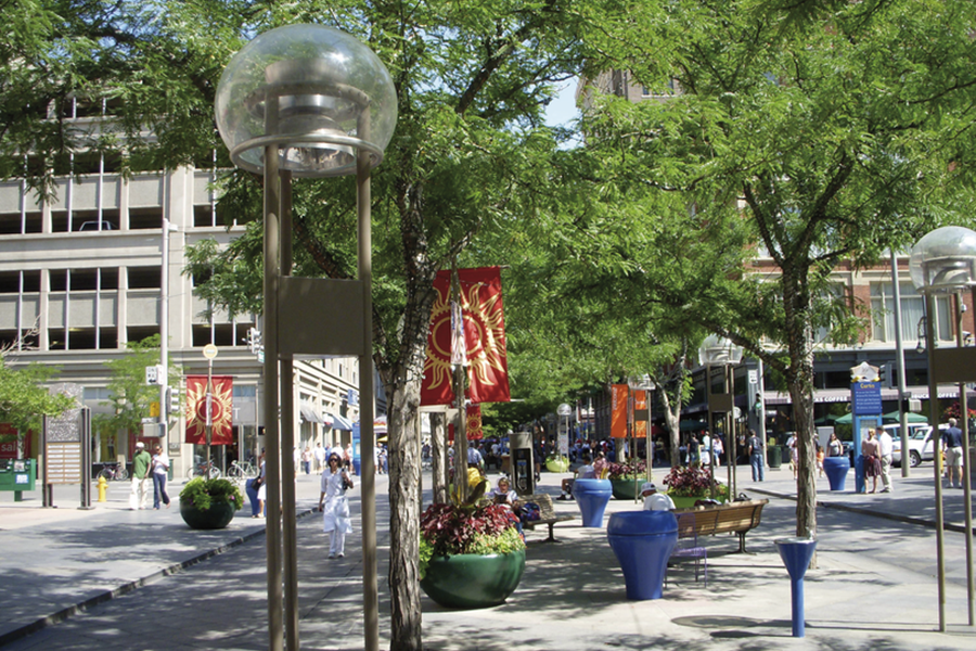 Ground-level view of a sunlit pedestrian mall lined with leafy green trees, modern globe street lamps, blue planters, and colorful sun-patterned banners.