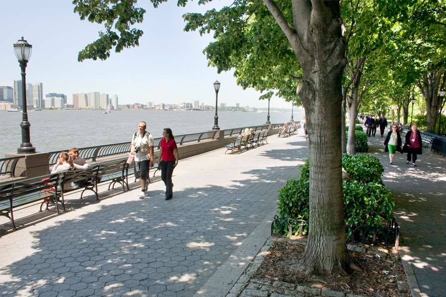 Pedestrians walk along a wide, paved riverside path lined with green trees, benches, and classic street lamps, with a city skyline visible across the water.