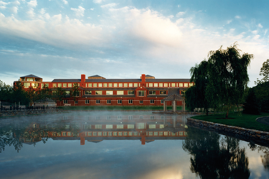 A wide shot of a long, red-brick building reflected in a misty, still pond at dawn. A small wooden gazebo sits on a stone pier at the water's edge.