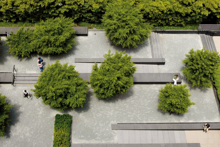 Bird's-eye view of a minimalist courtyard showing a pattern of dark stone benches and steps set against light gray gravel, interspersed with neatly manicured green trees.
