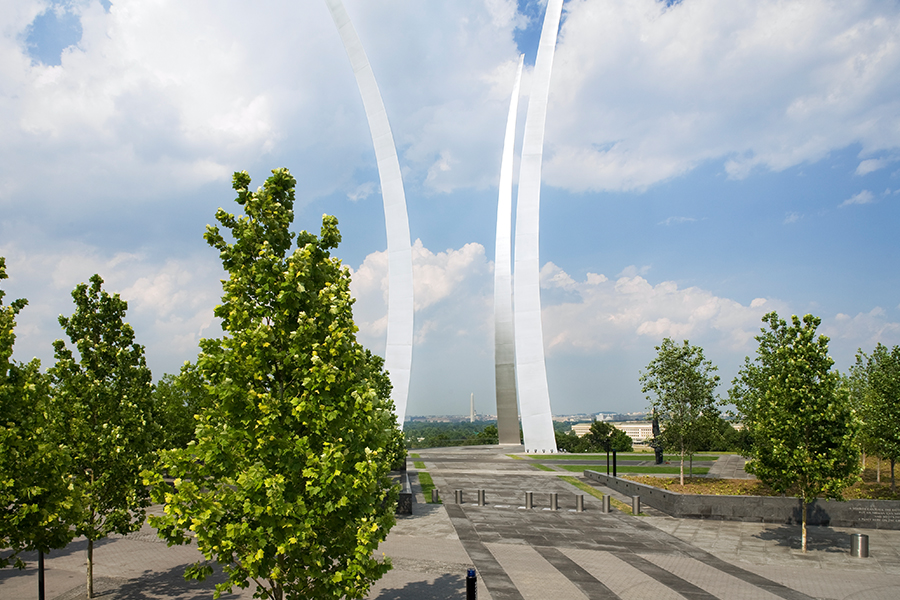 A paved plaza featuring vibrant green trees and towering, curved stainless steel spires reaching toward a cloudy blue sky. In the distant background, the Washington Monument is visible.