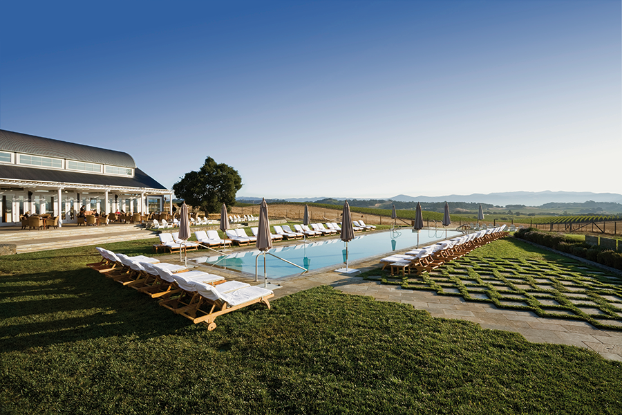 A wide shot of a luxury resort pool area featuring rows of wooden lounge chairs, a large barn-style pavilion, and a rolling vineyard landscape under a clear blue sky.