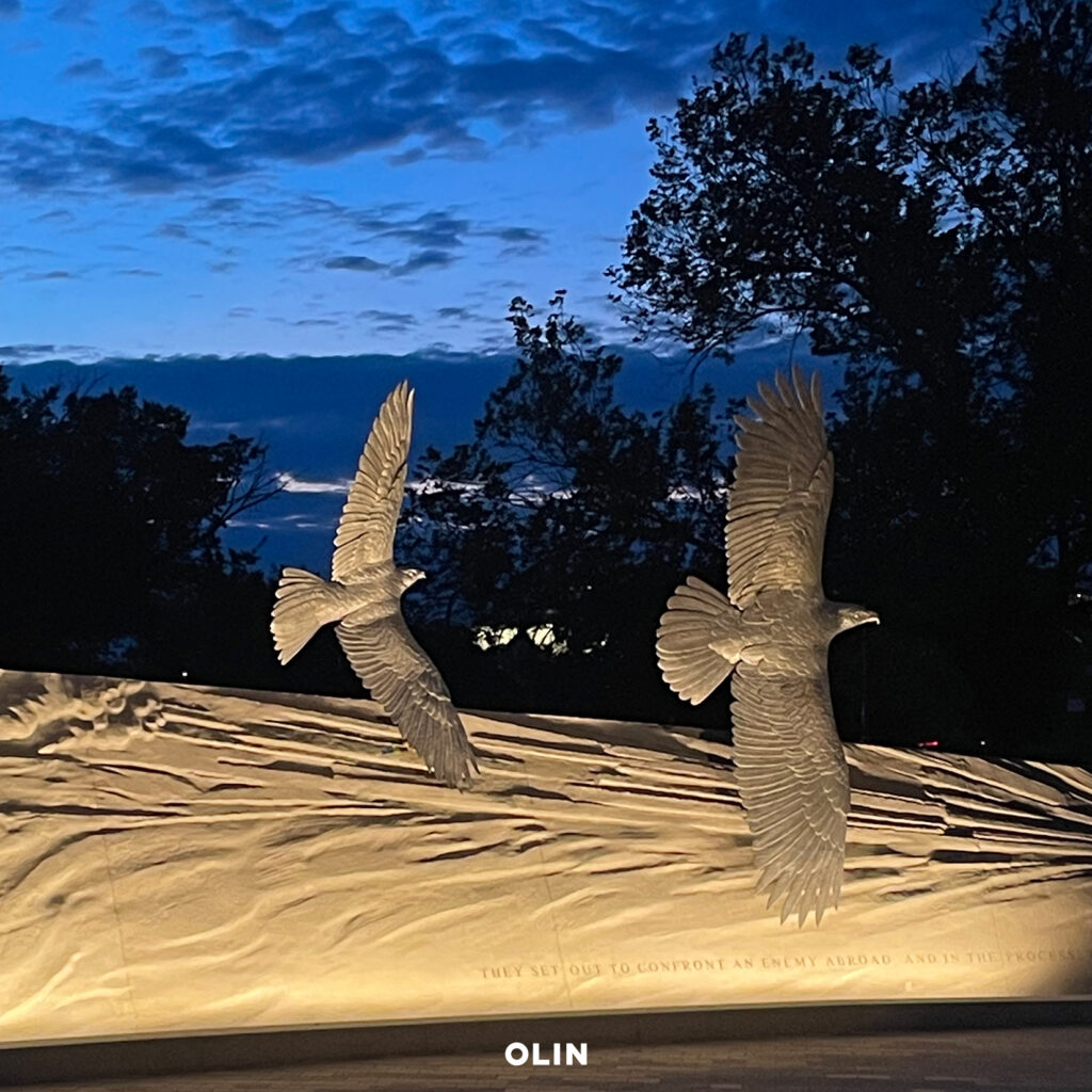 Illuminated falcon sculptures appearing to soar above an illuminated relief wall at dusk.