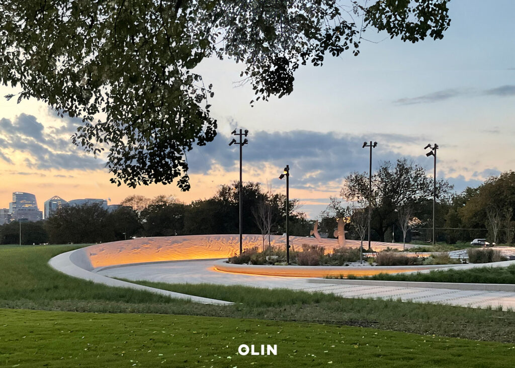 Wide view of the curved memorial plaza and softly lit earthwork wall at sunset.