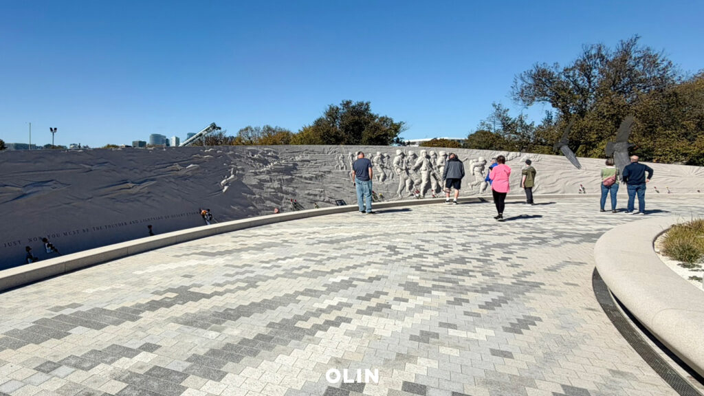Visitors walking along a curved stone plaza and viewing a bas-relief wall depicting soldiers in action.
