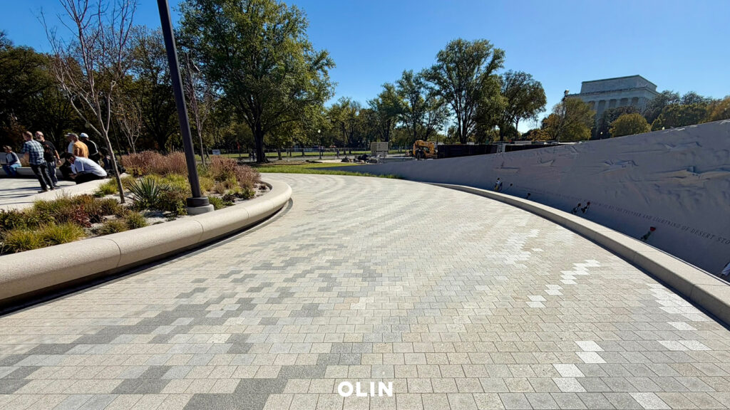 Paved pathway curving alongside a low inscribed memorial wall, with landscaped plantings and the Lincoln Memorial visible in the background.