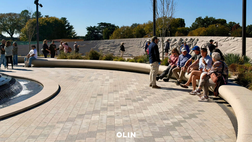Visitors seated on curved benches and walking around a circular fountain plaza near a sculpted relief wall on a sunny day.