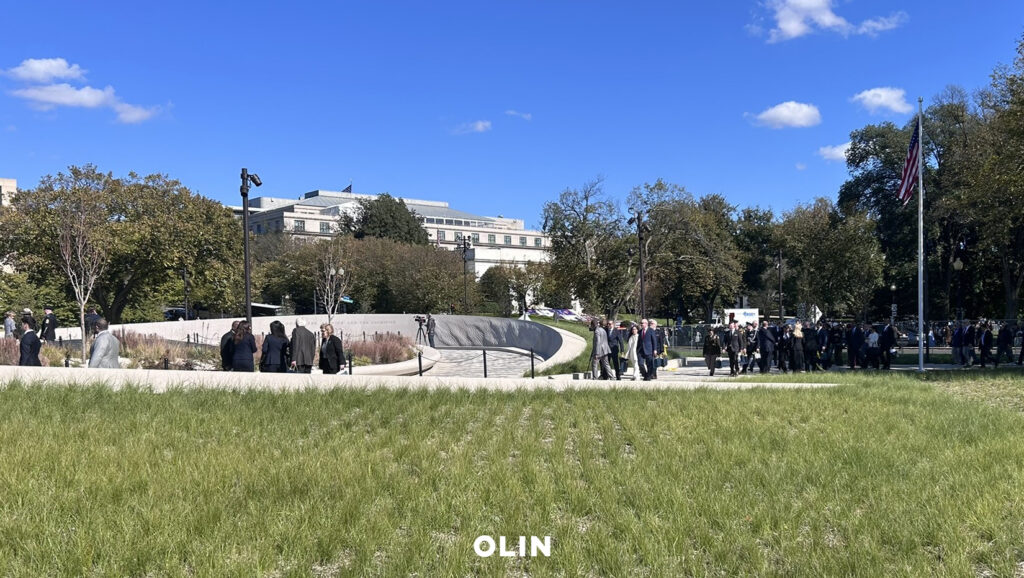 Attendees in formal attire gathering along the grassy grounds and curved entrance of the memorial site.