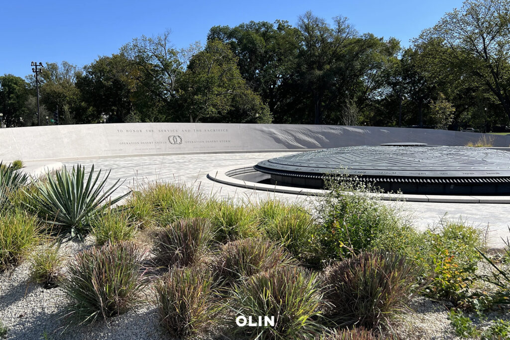A circular water feature and curved inscribed memorial wall set among native grasses and shrubs under a clear blue sky.