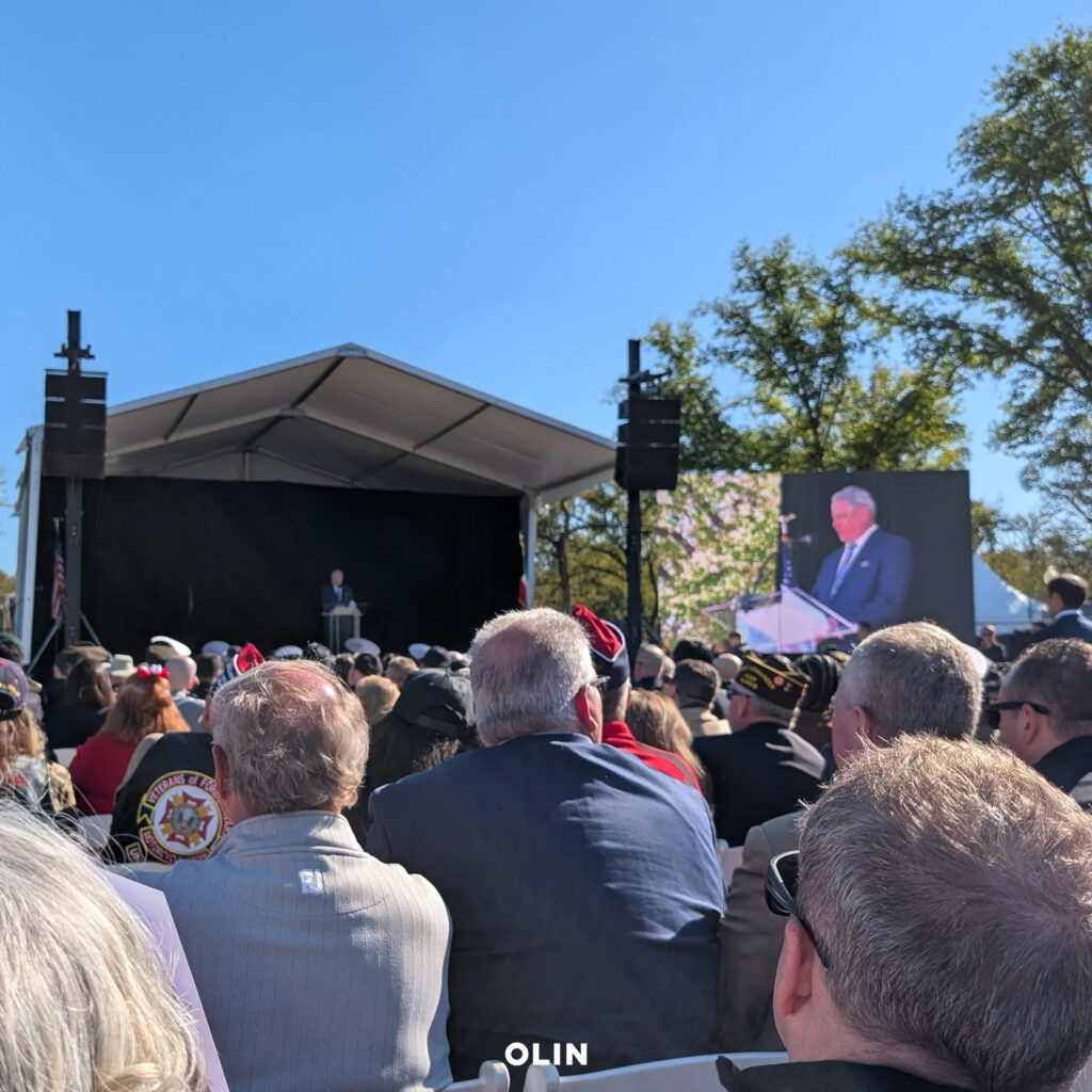 Audience seated outdoors facing an event stage and video screen during a dedication ceremony.