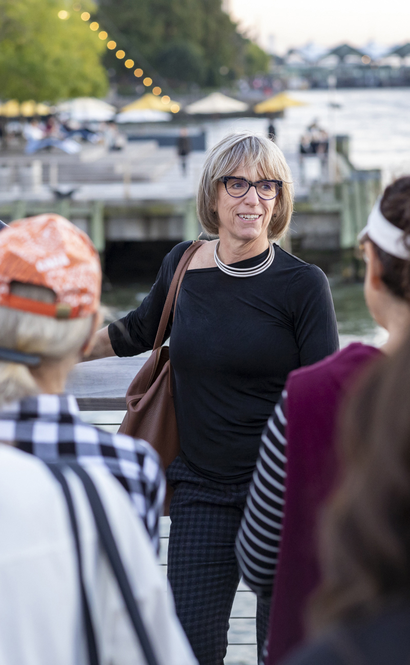 Lucinda Sanders, a smiling woman with blonde hair and glasses leading a project tour on an outdoor boardwalk by the water.