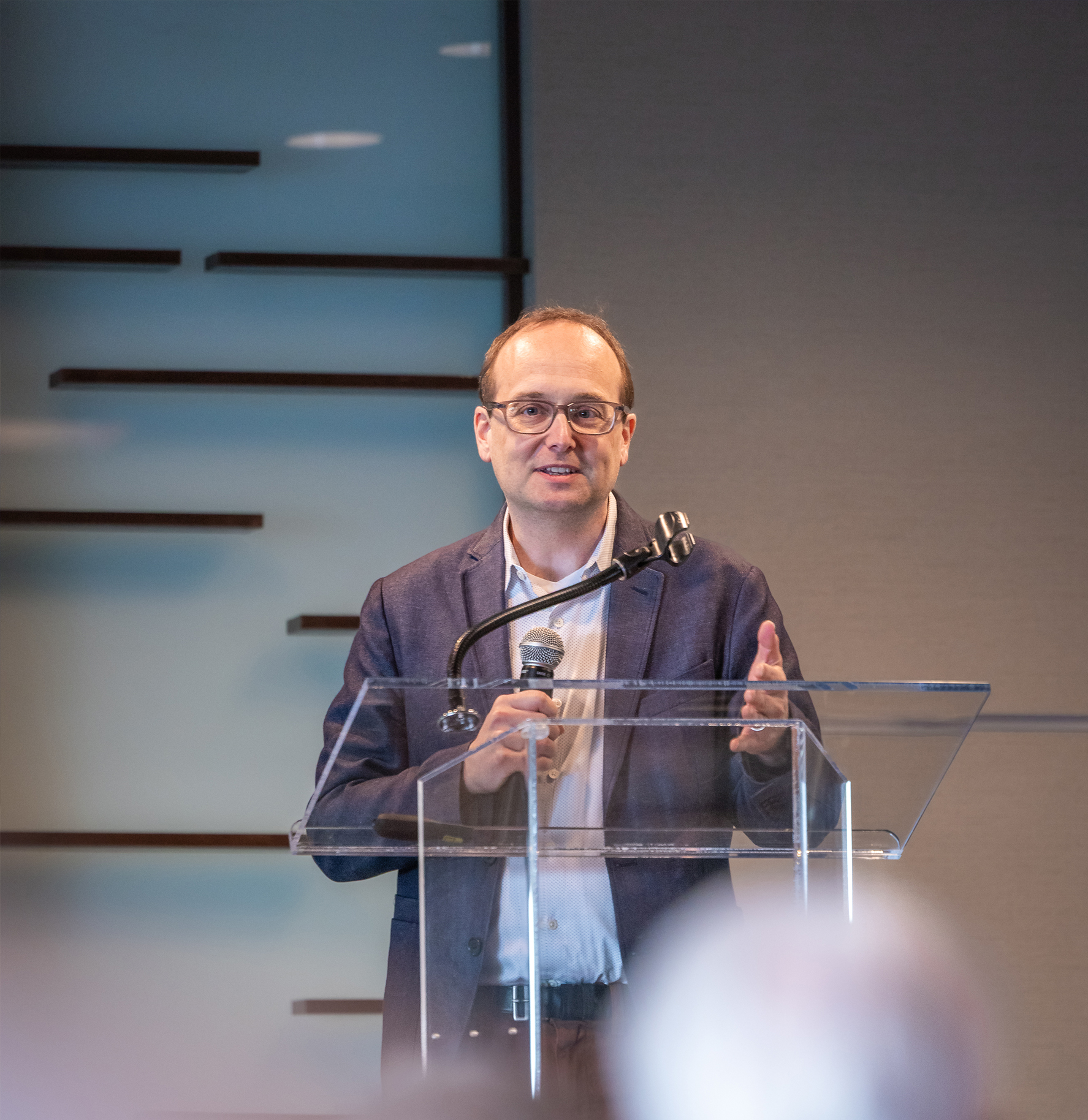 Richard Roark, a man in a blue blazer and glasses speaking at a transparent podium during a professional lecture.